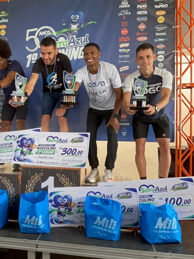 Kauê de Souza no pódio da 1ª Corrida Supermercados Serra Azul, com o troféu na mão, diante do painel dos 50 anos da rede